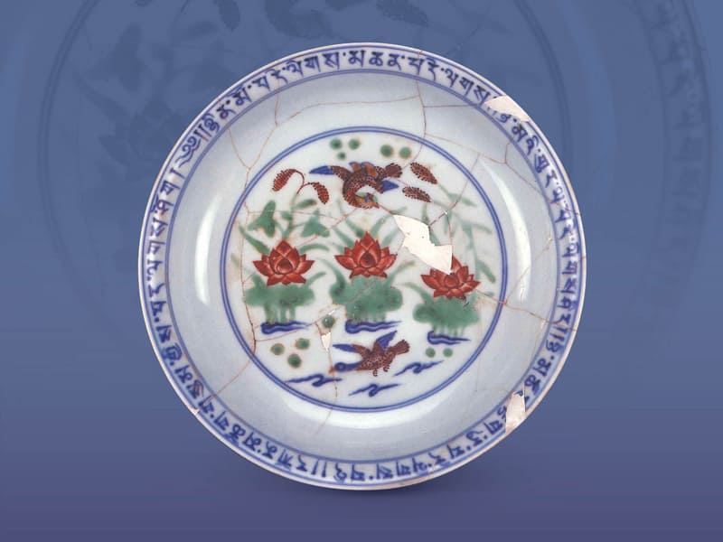 明宣德．五彩鴛鴦蓮池紋盤+藍色藏文、水波及邊框是釉下青花，其他的紅、綠、褐等紋飾則為釉上彩繪。（圖一）+圖片來源：《景德鎮珠山出土永樂宣德官窯瓷器展覽》，頁261，圖89。